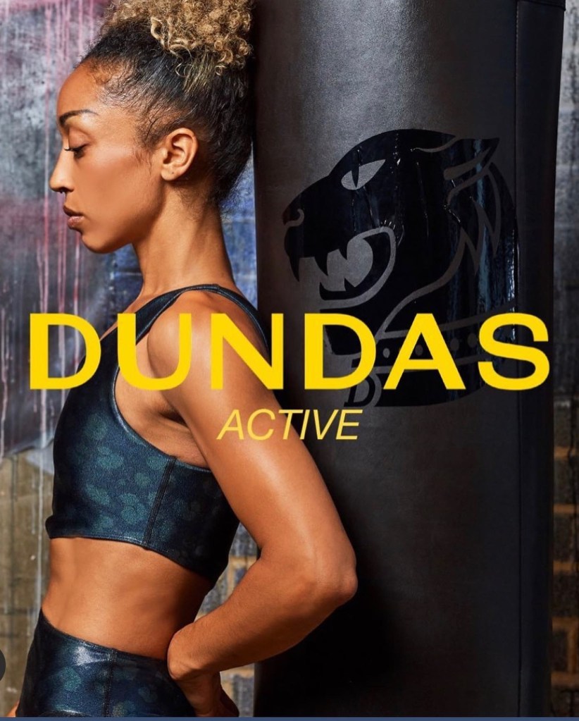 This is a sports active wear shoot for the fashion brand Dundas World. The model is standing with her back leaning up to a black punching bag. The yellow logo of Dundas World is running across the centre of the image.