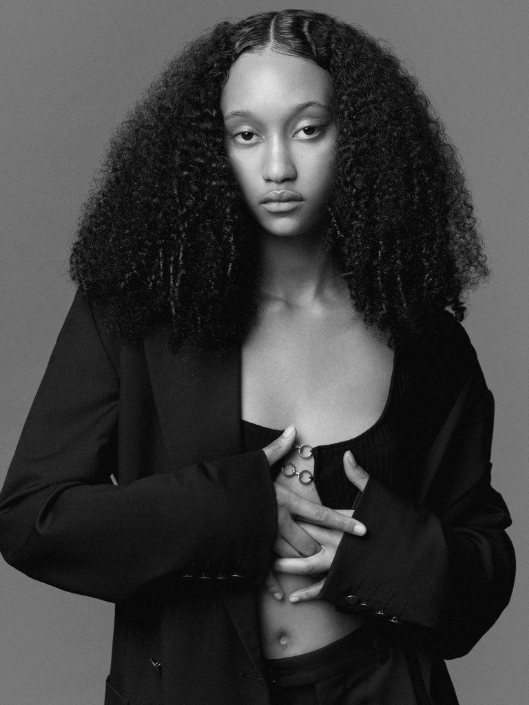 A black and white fashion shot taken for the modelling agency Present Models. The makeup is very natural with a flawless glowing looking skin. Her afro hair is parted down the centre and gelled at the parting. Her facial expression is strong and moody like. This image is cropped at the hips so you can see her skin and and hair beautifully. 